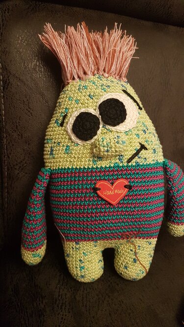 Gehäkeltes Amigurumi-Monster in Pastellgrün mit gestreiftem Torso, großen Augen und Fransenhaar