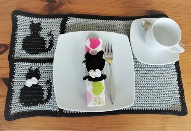 Gehäkeltes grau-schwarzes Platzset mit schwarzen Katzenmotiven und schwarzem Serviettenring, angerichtet mit Teller, Besteck und Tasse auf Holztisch.