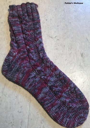 Gestrickte Socke mit Zopfmuster in Lila, Pink und Grau auf hellem Untergrund.