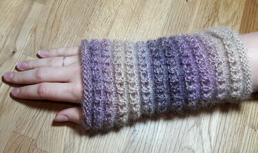 Gestrickte melierte Armstulpe in Lila-, Beige- und Grautönen an einem Handgelenk auf Holzfußboden.