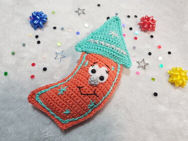Gehäkelte orangefarbene Deko-Rakete mit mintgrünem Hut und aufgesticktem Gesicht, umgeben von buntem Konfetti.
