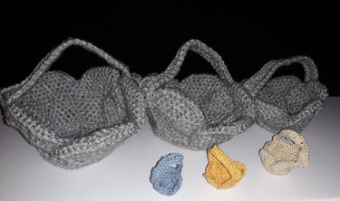 Gehäkelte Körbe in Grau mit Griffen, davor drei kleine gehäkelte Utensilos in verschiedenen Farben