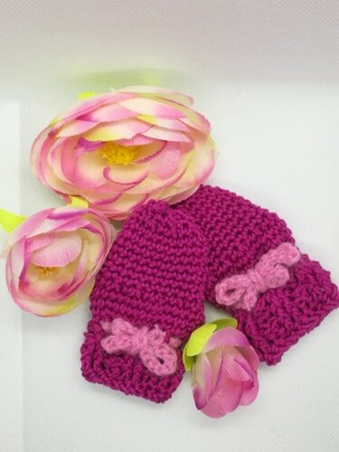 Zwei fuchsiafarbene gehäkelte Babyfäustlinge mit rosa Schleifen neben rosa Blumen