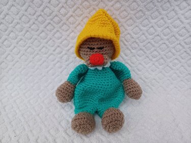 Gehäkelte Kuschelpuppe in Türkis mit gelber Zipfelmütze und roter Nase auf weißer, strukturierter Unterlage.