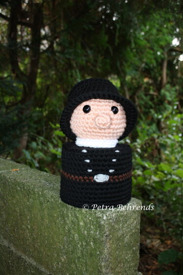 Gehäkelte Schornsteinfeger-Figur in schwarzer Kleidung auf moosbewachsener Steinmauer vor grünem Hintergrund