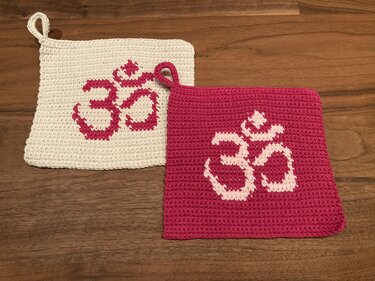 Zwei quadratische gehäkelte Topflappen auf Holztisch; weiß mit rosa Om und rosa mit weißem Om, beide mit Aufhängeschlaufe