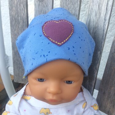 Babypuppe mit blauem Mützchen und lila Herz-Applikation mit gelber Ziernaht