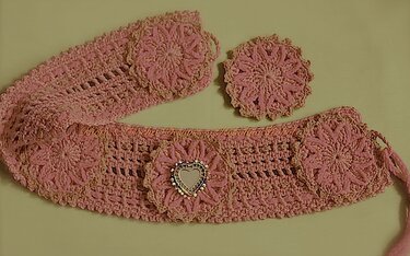 Rosa gehäkelter Gürtel mit drei Blumenapplikationen und herzförmigem Zierelement, daneben ein rundes gehäkeltes Blumenmotiv.