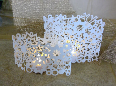 Zwei weiße Papier-Windlichter mit ausgeschnittenen Schneeflocken- und Sternmotiven, innen warm beleuchtet, vor goldenem Hintergrund.