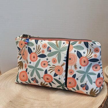 Kosmetiktasche mit Blumenmuster, Reißverschluss und Perlenanhänger auf Holzuntergrund.