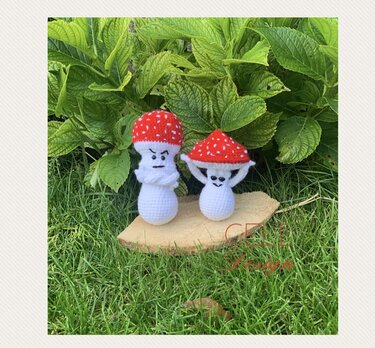 Zwei gehäkelte Pilze mit roten Kappen und weißen Punkten auf einer Holzscheibe vor grünen Blättern