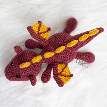 Gehäkelter Amigurumi‑Drache burgunderrot mit gelben Akzenten auf weißem, flauschigem Untergrund