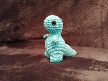 Türkis gehäkelter sitzender Amigurumi-Dinosaurier mit schwarzen Knopfaugen auf braunem Stoff