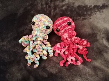 Zwei schnell gehäkelte Mini-Quallen mit gedrehten Tentakeln, pastell und rosa, auf dunklem Stoff