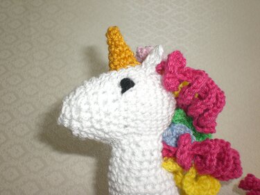 Weißes gehäkeltes Einhorn-Amigurumi in Seitenansicht mit gelbem Horn und bunter, lockiger Mähne