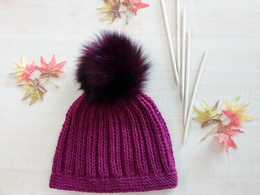 Lila Rippenstrickmütze mit dunklem Bommel auf hellem Holzuntergrund, daneben Stricknadeln und Herbstblätter.
