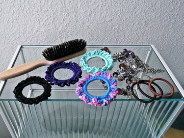 Holzbürste, mehrere bunte gehäkelte Haargummis und eine Kette auf einer Glasablage, Nahaufnahme.