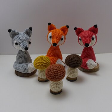 Drei gehäkelte Amigurumi-Füchse (grau, orange, rot) auf Holzscheiben mit vier Häkelpilzen im Vordergrund