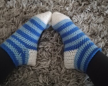 Paar Füße in blau-weiß gestreiften, gehäkelten Socken auf einem grauen, flauschigen Teppich.