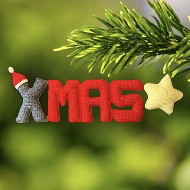 Gehäkelte Buchstaben 'XMAS' in Rot und Grau mit kleinem gelben Stern vor grünem Hintergrund