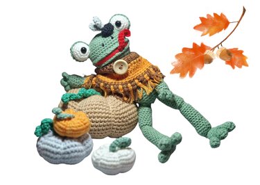 Grüner gehäkelter Amigurumi-Frosch mit Fliege und Poncho auf einem Kürbis, umgeben von kleineren Kürbissen