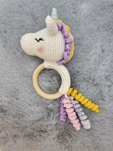 Gehäkelter weißer Einhorn-Greifring mit pastellfarbener Mähne und Holzring auf grauem Plüschuntergrund.