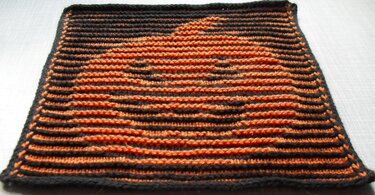 Gestricktes quadratisches Dekostück in Schwarz und Orange mit Kürbismotiv auf hellem Untergrund