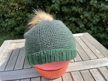 Dunkelgrüne gestrickte Mütze mit braunem Fellbommel auf einem Schaummodel, auf einem hölzernen Tisch