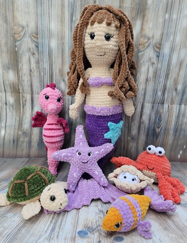 Gehäkelte Meerjungfrau mit braunen Locken und lila Schwanz, umgeben von gehäkelten Meerestieren wie Seepferdchen, Seestern, Schildkröte, Krabbe, Muschel und Fisch.