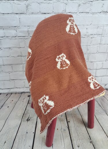 Reddish-brown knitted baby blanket with white owl motifs draped over a stool