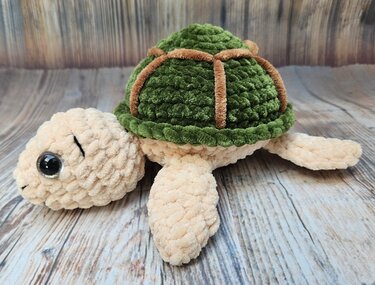 Gehäkelte Amigurumi‑Schildkröte mit grünem Panzer und beigem Körper auf Holzboden.