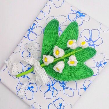 Nahaufnahme einer gehäkelten Maiglöckchen-Applikation mit grünen Blättern, weißen Glockenblüten und weißem Spitzenband auf gemustertem Papier.