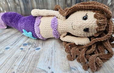 Gehäkelte Meerjungfrau-Amigurumi mit lila Schwanz und braunen Locken auf Holzuntergrund