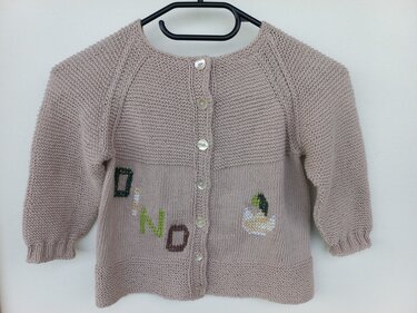Beige gestrickte Babyjacke mit DINO-Stickerei und Dinosaurier-Ei, an einem Kleiderbügel