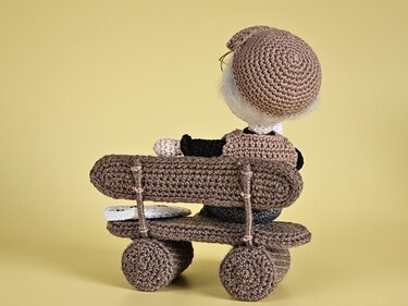 Gehäkelte Amigurumi-Puppe sitzt von hinten auf einer braunen Bank mit Rädern vor gelbem Hintergrund.