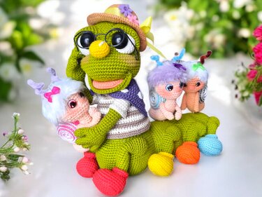 Grüne gehäkelte Schnecke mit Hut und Brille, die drei kleine gehäkelte Schneckenbabys trägt, vor unscharfem Blumenhintergrund.