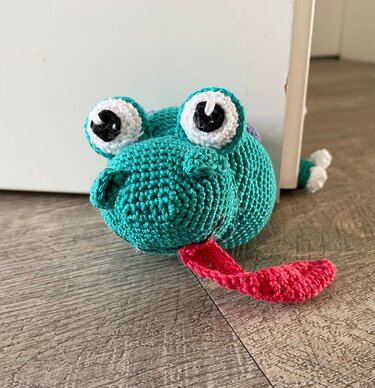 Gehäkelter türkisfarbener Drache mit großen Augen und roter Zunge, der als Türstopper eine weiße Tür offen hält.