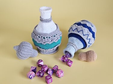 Zwei gehäkelte Behälter, ein aufrecht in Weiß und Türkis, ein liegend in Blau und Grau, mit violetten Spielwürfeln vor gelbem Hintergrund.