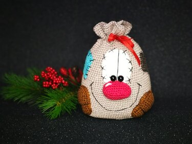 Beigefarbener gehäkelter Geschenksack mit fröhlichem Gesicht, rosa Nase und roter Schleife neben Tannengrün und roten Beeren.