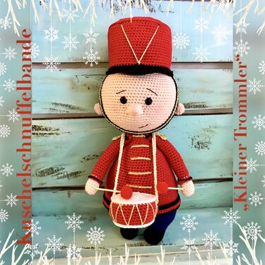 Gehäkelte Amigurumi-Puppe als kleiner Trommler in roter Uniform mit Trommel vor hellem Holzbrett