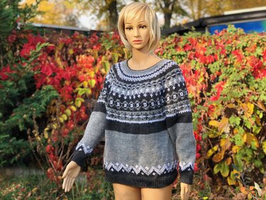 Schaufensterpuppe trägt gestrickten Damenpullover mit Fair-Isle-Muster in Grau, Schwarz und Weiß vor herbstlichem Pflanzenhintergrund