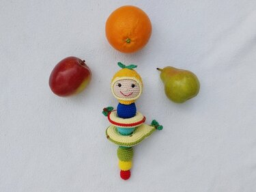 Gehäkelte bunte Stapelraupe mit lächelndem Gesicht neben Apfel, Birne und Orange auf weißem Hintergrund.