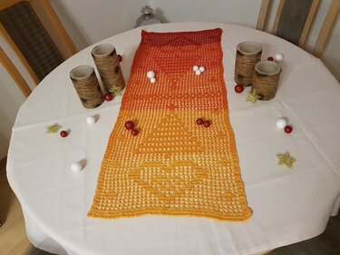 Gehäkelter Tischläufer in Gelb‑Orange‑Rot mit Lochmuster auf rundem weißen Tisch, dekoriert mit Kerzenhaltern und Kugeln