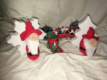 Zwei gehäkelte rote Weihnachtswichtel mit weißen Bärten, flankiert von weißen Holzfiguren (Schneeflocke, Tannenbaum) und einer Spielzeugeisenbahn im Hintergrund.