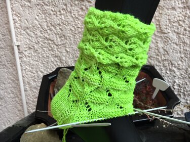 Nahaufnahme einer unfertigen neongrünen Socke mit Loch- und Spitzenmuster sowie Rüschenrand auf Stricknadeln.