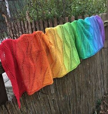 Gestrickter Schal in Regenbogenfarben mit Lochmuster, über einem Gartenzaun drapiert