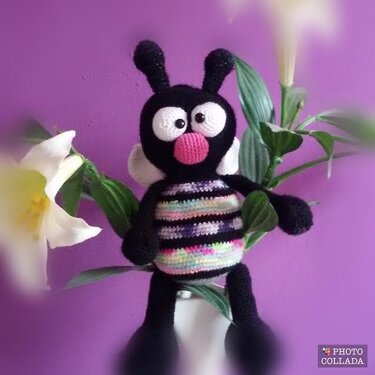 Gehäkelte schwarze Amigurumi-Biene mit buntem gestreiftem Bauch und weißen Flügeln vor lila Hintergrund