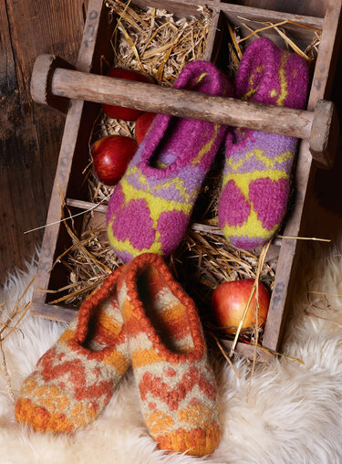 Zwei Paar gemusterte Hausschuhe, eines auf Schaffell, eines in einer Holzkiste mit Stroh und Äpfeln