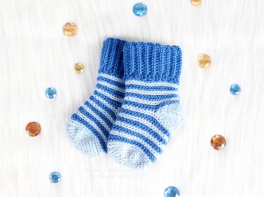 Blue and white striped crocheted baby socks on a soft white background with scattered gems.