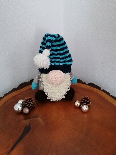 Gehäkelter Wichtel mit blau-weiß gestreifter Mütze, weißem Bommel und flauschigem Bart auf einer Holzscheibe mit Weihnachtskugeln und Tannenzapfen.